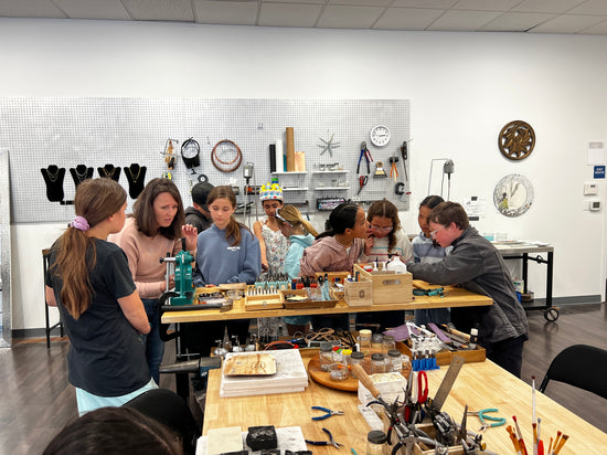 Group of people in a workshop setting making jewelry with tools and materials on tables.