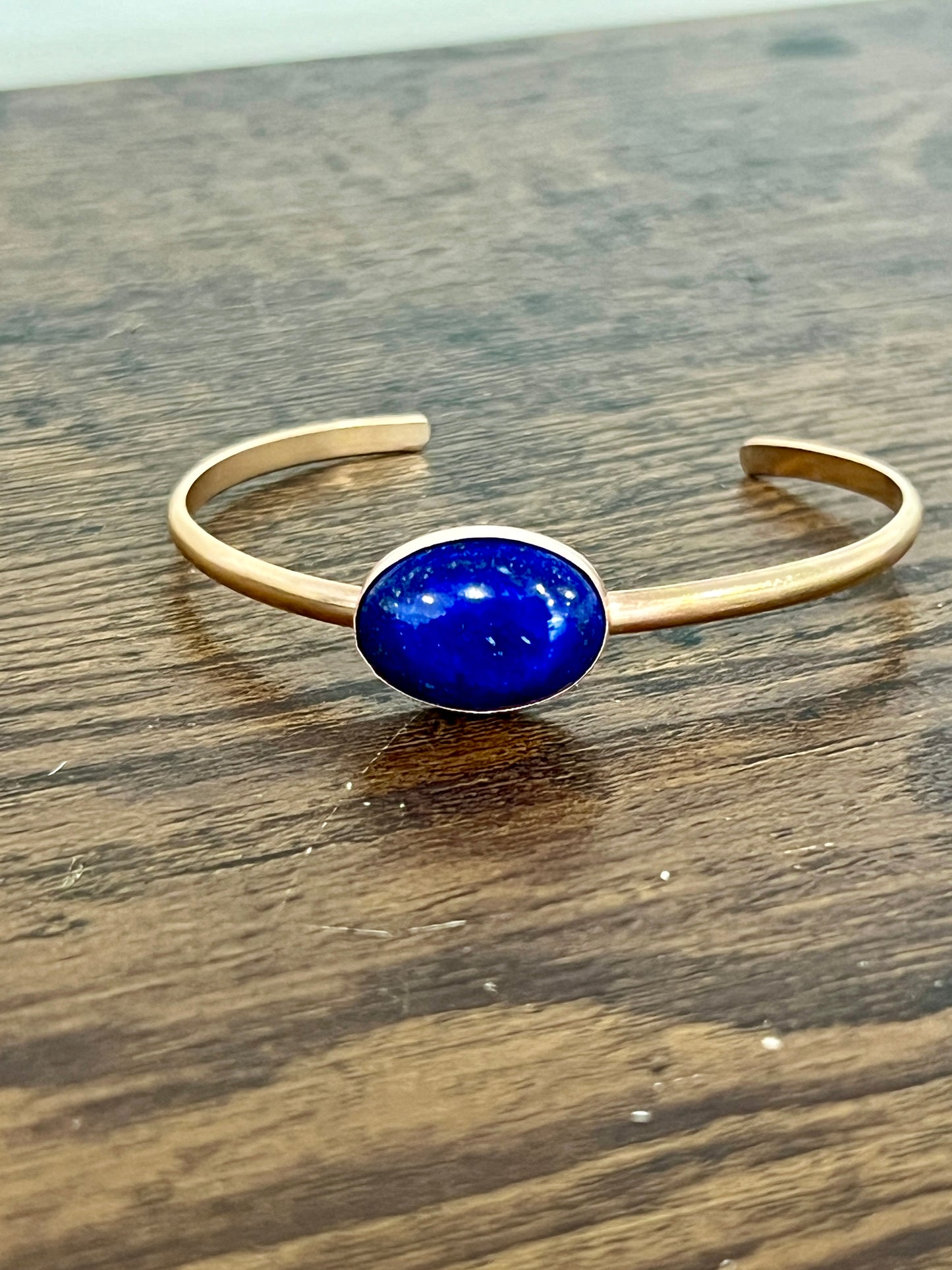 A lapis lazuli stone with gold flecks set on a 14k gold fill cuff bracelet on a wooden surface.